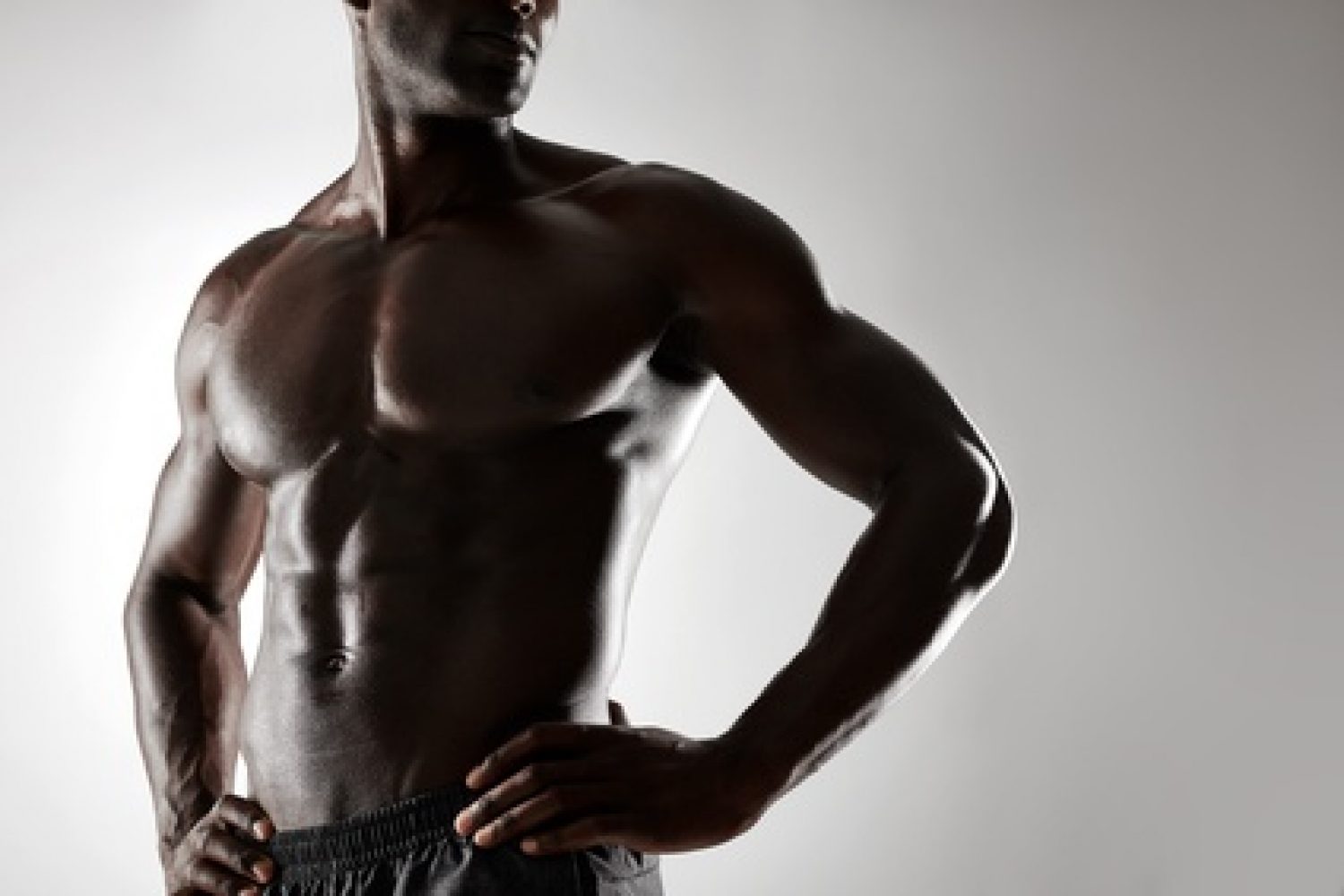 Cropped shot of young man with muscular body standing against grey background. Shirtless male model with hands on hips.
