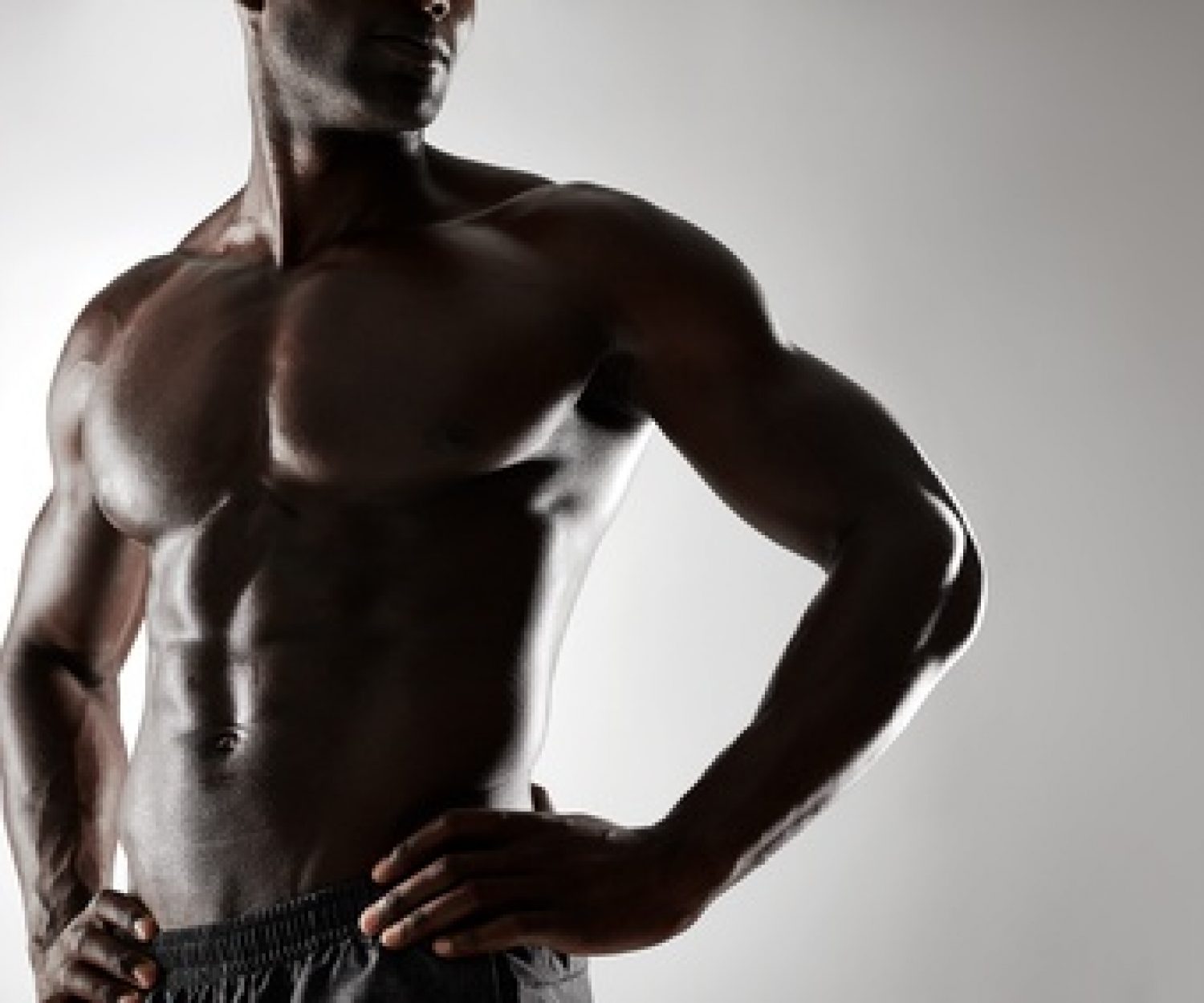 Cropped shot of young man with muscular body standing against grey background. Shirtless male model with hands on hips.